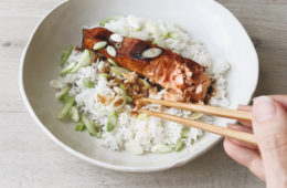 Mirin glazed salmon on rice in a white bowl with a hand holding chopsticks reaching in