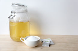 Kombucha in a large jar with sugar in a bowl and green tea bags on the side