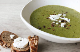 Pea and Watercress soup in a white bowl with a rye bagel and feta cheese on a wooden surface