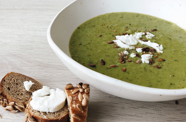 Pea and Watercress soup in a white bowl with a rye bagel and feta cheese on a wooden surface